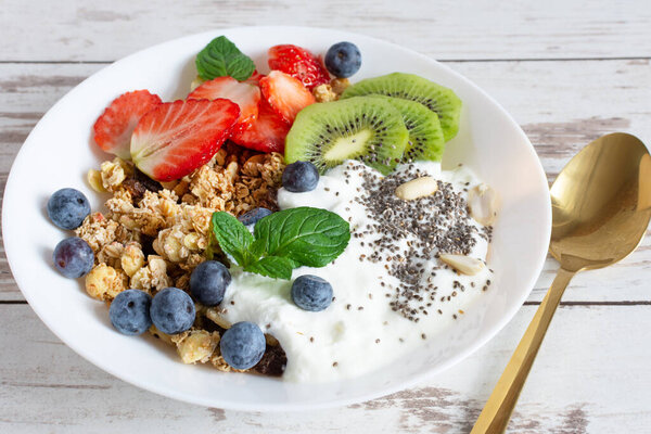 White ceramic bowl of homemade granola with yogurt, fresh berries and fruits on white wooden table background. Healthy eating concept.