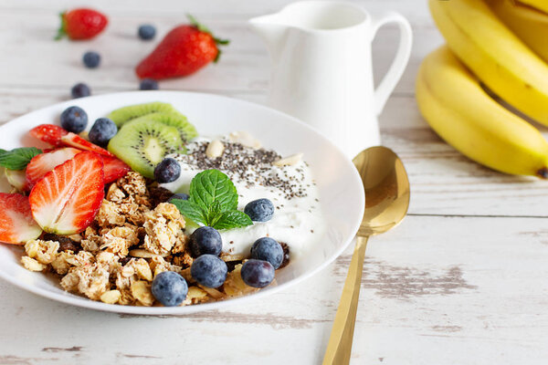 White ceramic bowl of homemade granola with yogurt, fresh berries and fruits on white wooden table background. Healthy eating concept.