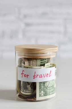 Money savings for travel in a glass jar on white wood table over white brick wall. Savings for travel budget concept. clipart