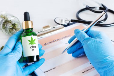 Doktor hasta için CBD yağı yazıyor. Bir şişe Cbd yağı, kenevir tohumu ve kuru yapraklar beyaz ahşap masadaki tıbbi reçete kağıdının üstünde. Tıbbi marihuana, alternatif tıp konsepti.