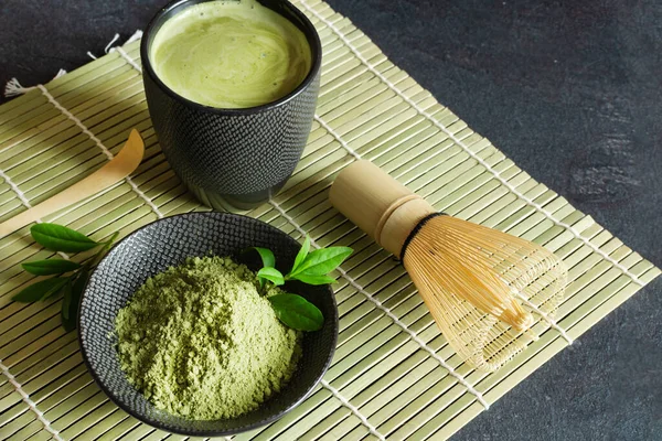 Organic green matcha tea and tea accessories on japanese mat on black background. Japanese tea ceremony concept. Chashaku spoon and chasen bamboo whisk for brewing matcha tea.