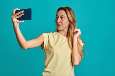 Mavi arka planda yarım boy telefonları olan güzel bir kız portresi. Genç bir kadın selfie çekiyor ve telefonda konuşuyor..