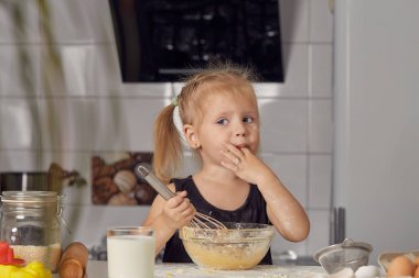 Küçük kız, un, süt ve modern bir mutfaktaki bir masada oturan tatlı bir pasta hazırlıyor. Kız çırpı tutuyor, yumurta karıştırıyor, krep hamuru hazırlıyor. Küçük aşçı.