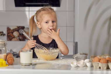 Küçük kız, un, süt ve modern bir mutfaktaki bir masada oturan tatlı bir pasta hazırlıyor. Kız çırpı tutuyor, yumurta karıştırıyor, krep hamuru hazırlıyor. Küçük aşçı.