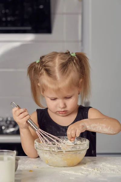 Little little girl prepares a sweet cake with flour, milk, sitting on chairs at a table in a modern kitchen. Girl holds a whisk, stirring eggs in a bowl, prepares pancake dough. Little cook.