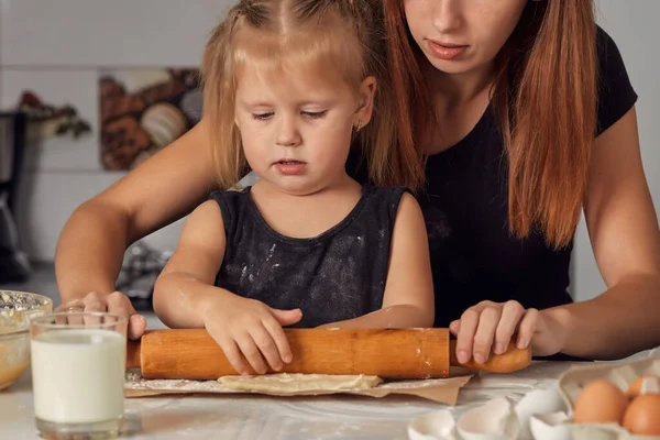 Mother and daughter preparing a sweet cake using flour, milk, sitting on chairs at a table in a modern kitchen. Girl holding a whisk, stirring eggs in a bowl, preparing pancake dough with her mom.