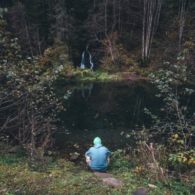 Adam bir şelale önünde bir göl kıyısında oturur.