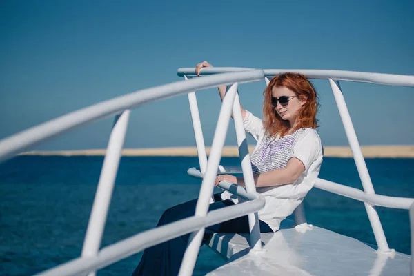Beautiful ginger woman in sun glasses sits on a white yacht in a Red sea with clear turquoise water. Relaxation at summer vacation under a sun.