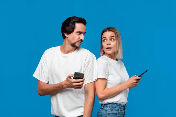 Femme blonde et brunet homme en tess blanche et jeans bleus regardant les écrans de leurs téléphones mobiles debout avec le dos à l'autre . — Photo