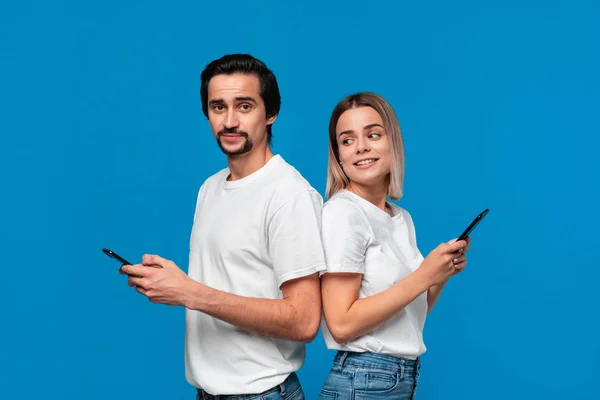 Femme blonde et brunet homme en tess blanche et jeans bleus regardant les écrans de leurs téléphones mobiles debout avec le dos à l'autre . — Photo