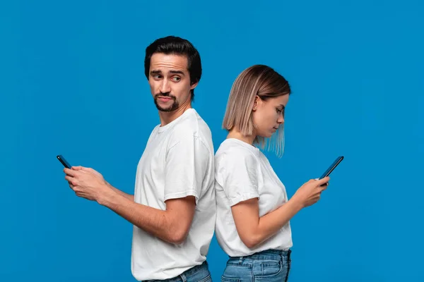 Femme blonde et brunet homme en tee-shirts blancs et jeans bleus avec téléphones mobiles debout isolé sur fond bleu. L'homme essaie de voir ce qu'il y a sur un écran de son freind . — Photo