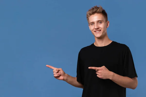 Jovem bonito em um tee preto mostrando com os dedos deixados no espaço vazio para publicidade . — Fotografia de Stock