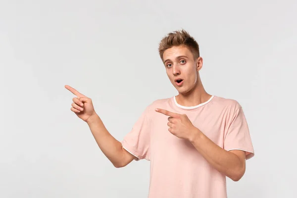Przystojny młody człowiek w różowej koszulce pokazuje z palcami w pustej przestrzeni na reklamę. — Zdjęcie stockowe