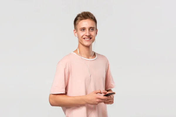 Улыбающийся блондин-подросток в розовой футболке слушает музыку на смартфоне в проводных наушниках и смотрит на камеру, изолированную на светло-сером фоне . — стоковое фото