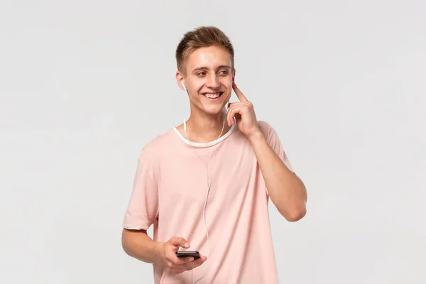 Leende blond tonåring i en rosa tee lyssna musik på en smartphone med trådbundna hörlurar och titta på kameran isolerad över ljusgrå bakgrund. — Stockfoto
