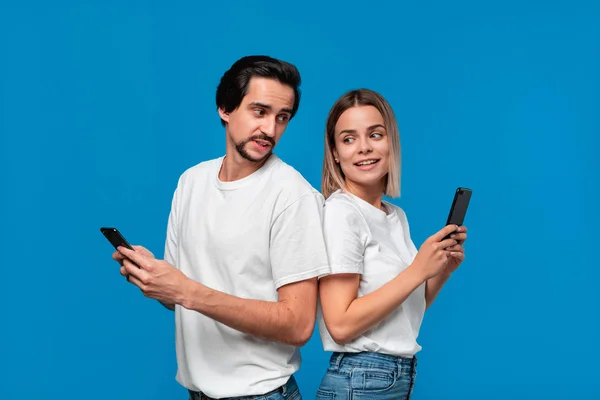 Femme blonde et brunet homme en tee-shirts blancs et jeans bleus regardant les écrans de leurs téléphones mobiles debout avec le dos à l'autre — Photo