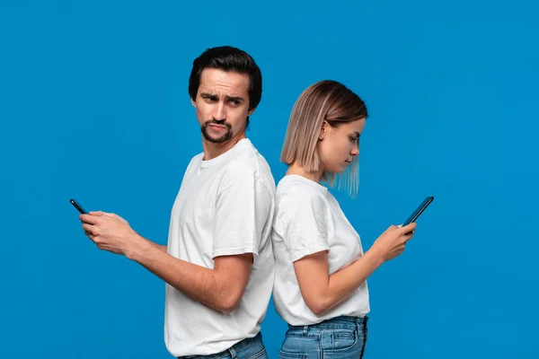 Femme blonde et brunet homme en tee-shirts blancs et jeans bleus avec téléphones mobiles debout isolé sur fond bleu. L'homme essaie de voir ce qu'il y a sur un écran de son freind . — Photo