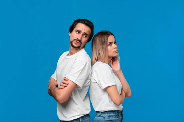 Upset couple of a young blond woman and brunet bearded man with mustaches in white t-shirts and blue jeans posing isolated over blue background.