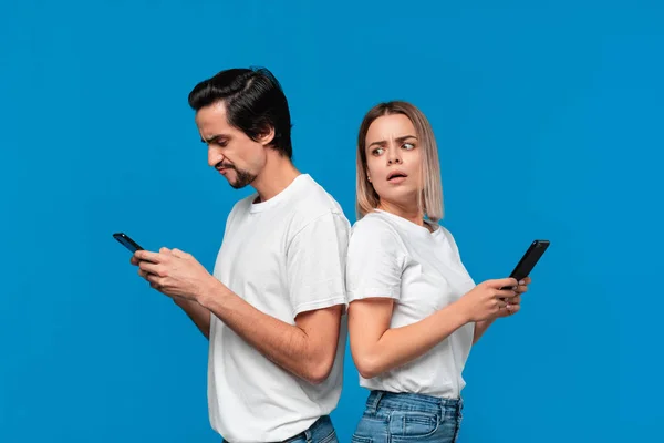 Femme blonde et brunet homme en tee-shirts blancs et jeans bleus avec téléphones mobiles debout isolé sur fond bleu. Fille essaie de voir ce qui est sur un écran de son partenaire . — Photo