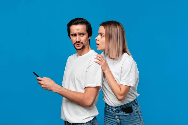 Femme blonde et brunet homme en tee-shirts blancs et jeans bleus avec téléphones mobiles debout isolé sur fond bleu. Fille essaie de voir ce qui est sur un écran de son partenaire . — Photo