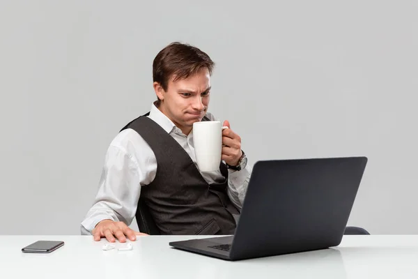 Empresario en una camiseta blanca y chaleco gris se sorprende comprobando la información en el ordenador portátil sentado en la mesa blanca bebiendo café . — Foto de Stock