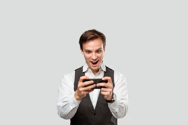 Joven guapo con camisa blanca y chaleco gris jugando en un smartphone . — Foto de Stock