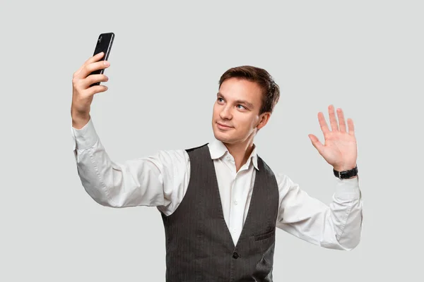 Joven hombre guapo en una camisa blanca y chaleco gris haciendo selfie con un teléfono móvil . — Foto de Stock