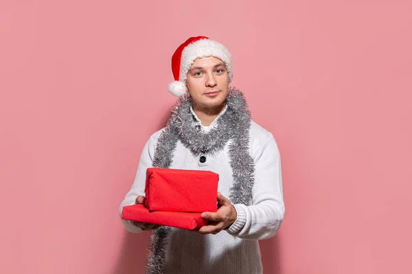 Bonito homem em um suéter branco e chapéu de Papai Noel vermelho segurando presentes de Natal em caixas vermelhas isoladas no fundo rosa . — Fotografia de Stock