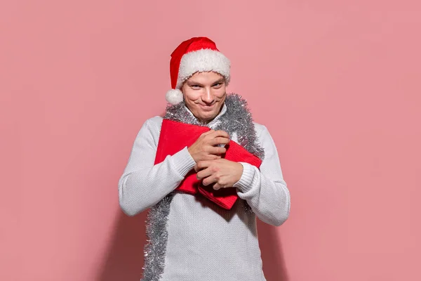 Fešák muž v bílém svetru a červený Santa klobouk drží pevně vánoční dárky v červené krabice izolované na růžovém pozadí. — Stock fotografie