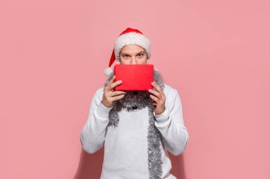 Beyaz kazaklı ve kırmızı şapkalı bir adam pembe arka planda izole edilmiş kırmızı kutularda Noel hediyeleri taşıyor..