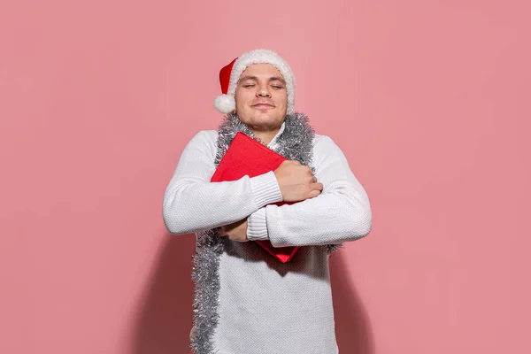 Bonito homem em um suéter branco e chapéu vermelho de Papai Noel segurando firmemente presentes de Natal em caixas vermelhas isoladas no fundo rosa. Feliz Natal e feliz Ano Novo — Fotografia de Stock