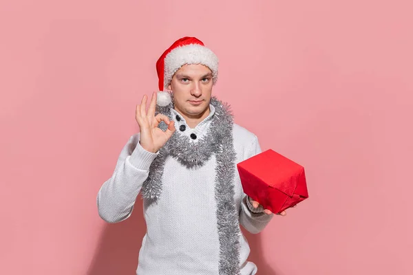 Muž v bílém svetru a červený Santa klobouk drží vánoční dárky v červených krabicích izolovaných na růžovém pozadí. — Stock fotografie