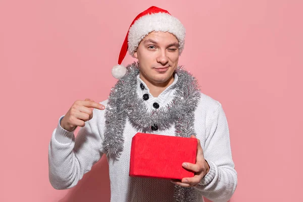 Muž v bílém svetru a červený klobouk Santa ukazuje na červenou krabici s vánoční dárky izolované na růžovém pozadí. — Stock fotografie