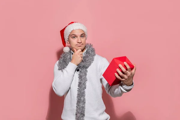 Homem de camisola branca e chapéu vermelho de Papai Noel olhando para a caixa vermelha para descobrir quais presentes estão em . — Fotografia de Stock