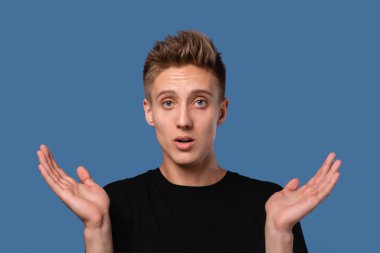 Şok olmuş siyah tişörtlü genç adam kayıp. Mavi arka planda izole edilmiş kameraya bakıyor..