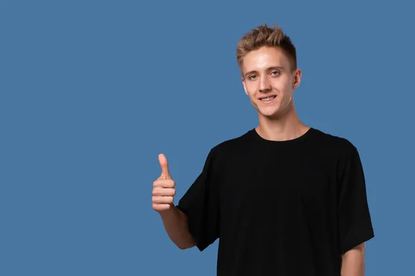 Sorrindo jovem bonito em uma camiseta preta segurando o polegar para cima mostrando sua avaliação . — Fotografia de Stock