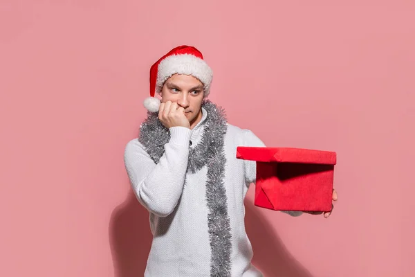 Hombre en un suéter blanco y sombrero rojo de Santa mirando la caja roja para averiguar qué regalos están en . — Foto de Stock