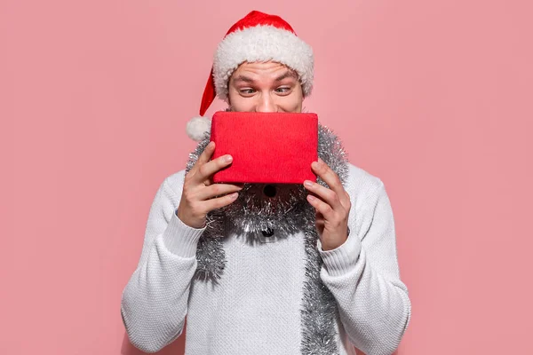 Homem de camisola branca e chapéu vermelho de Papai Noel escondido atrás da caixa vermelha com presentes de Natal isolados no fundo rosa . — Fotografia de Stock