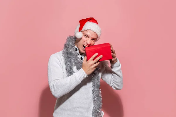 Férfi egy fehér pulóver és piros Mikulás kalap harapás karácsonyi ajándékok egy piros doboz elszigetelt rózsaszín háttér. — Stock Fotó