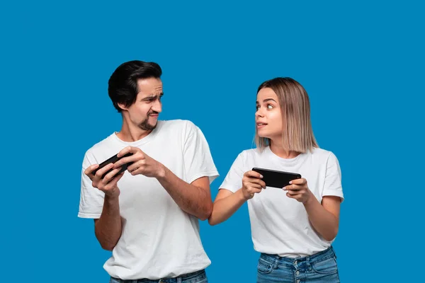 Par bär vita t-shirts stående isolerade över blå bakgrund, spela spel på mobiltelefon — Stockfoto