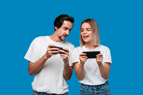 Couple portant des t-shirts blancs debout isolé sur fond bleu, jouer à des jeux sur téléphone mobile — Photo
