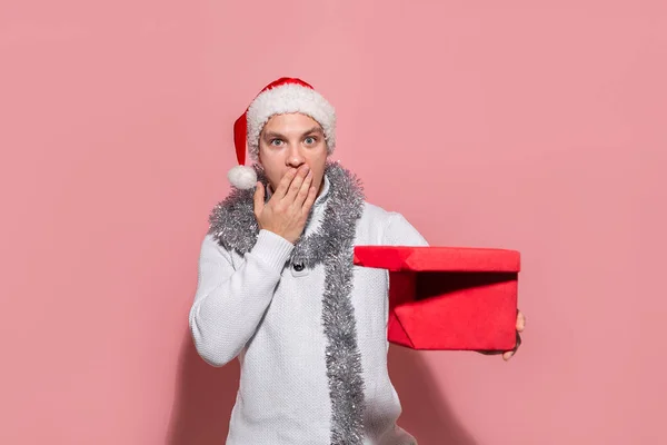 Hombre en un suéter blanco y sombrero rojo de Santa mirando la caja roja para averiguar qué regalos están en . — Foto de Stock