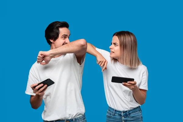 Pareja vistiendo camisetas blancas de pie aisladas sobre fondo azul, jugando juegos en el teléfono móvil — Foto de Stock