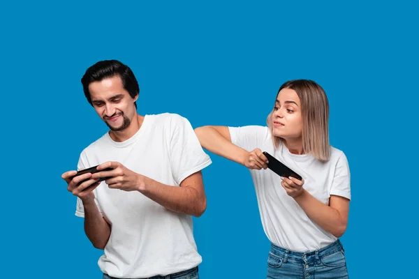 Pareja vistiendo camisetas blancas de pie aisladas sobre fondo azul, jugando juegos en el teléfono móvil — Foto de Stock