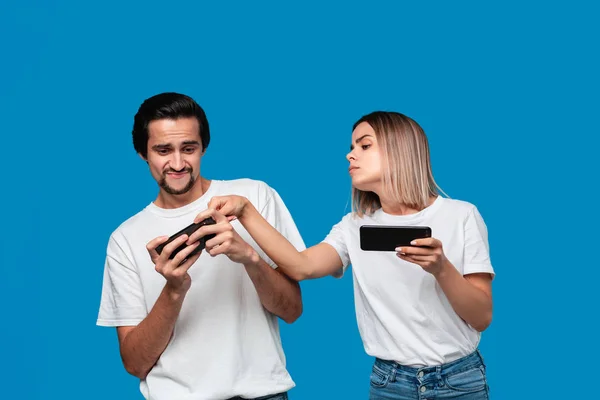 Para ubrana w białe koszulki stojąca na niebieskim tle, grająca w gry na telefonie komórkowym — Zdjęcie stockowe
