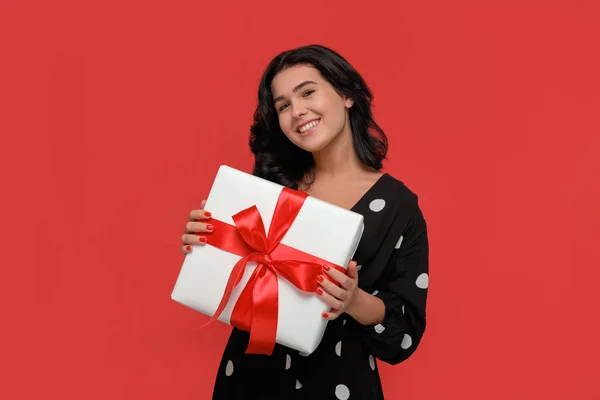 Brunette vrouw in een zwarte jurk glimlachen met kerst geschenkdoos van witte kleur met rood lint. — Stockfoto