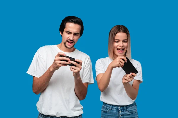 青い背景に白いTシャツを着たカップルが孤立し、携帯電話でゲームをプレイ ロイヤリティフリーのストック写真