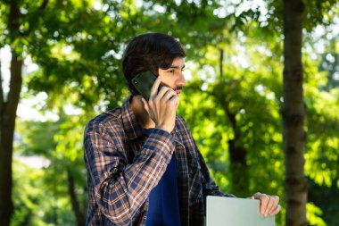 Parktaki akıllı telefondan konuşan dizüstü bilgisayarı olan genç bir serbest yazar..