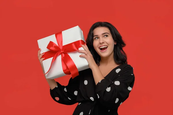 Brunetka kobieta w czarnej sukience uśmiechnięta gospodarstwa Christmas Giftbox w kolorze białym z czerwoną wstążką. — Zdjęcie stockowe