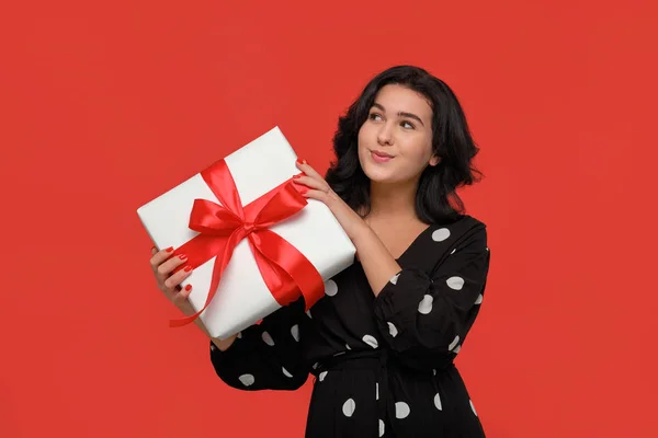 Mujer morena en un vestido negro sonriendo sosteniendo la caja de regalo de Navidad de color blanco con cinta roja . —  Fotos de Stock